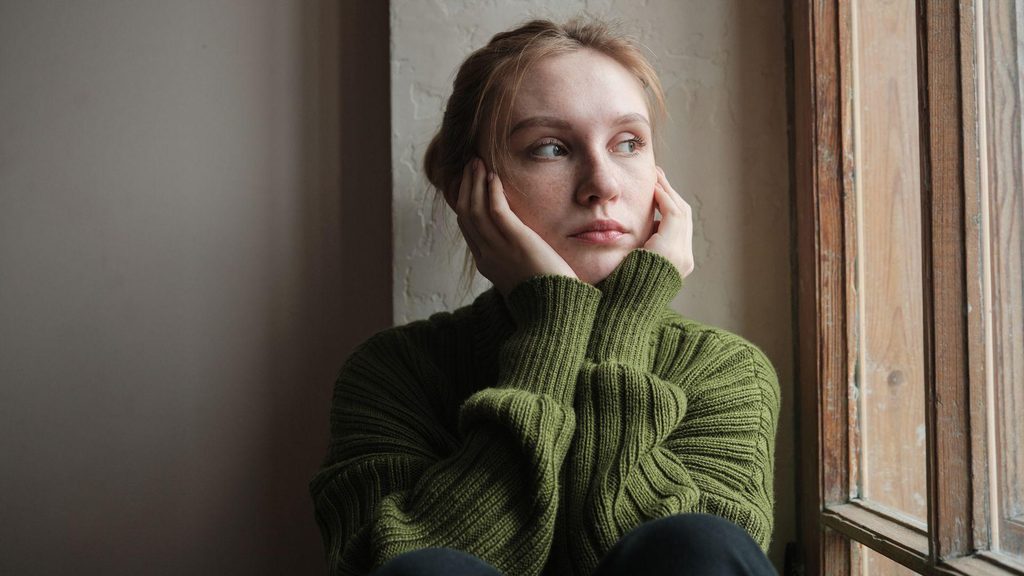Jeune femme en pull vert, pensive près d'une fenêtre, illustrant la peur du regard des autres.