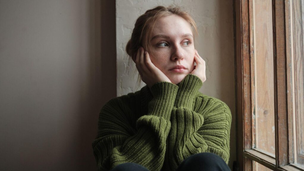Jeune femme en pull vert, pensive près d'une fenêtre, illustrant la peur du regard des autres.